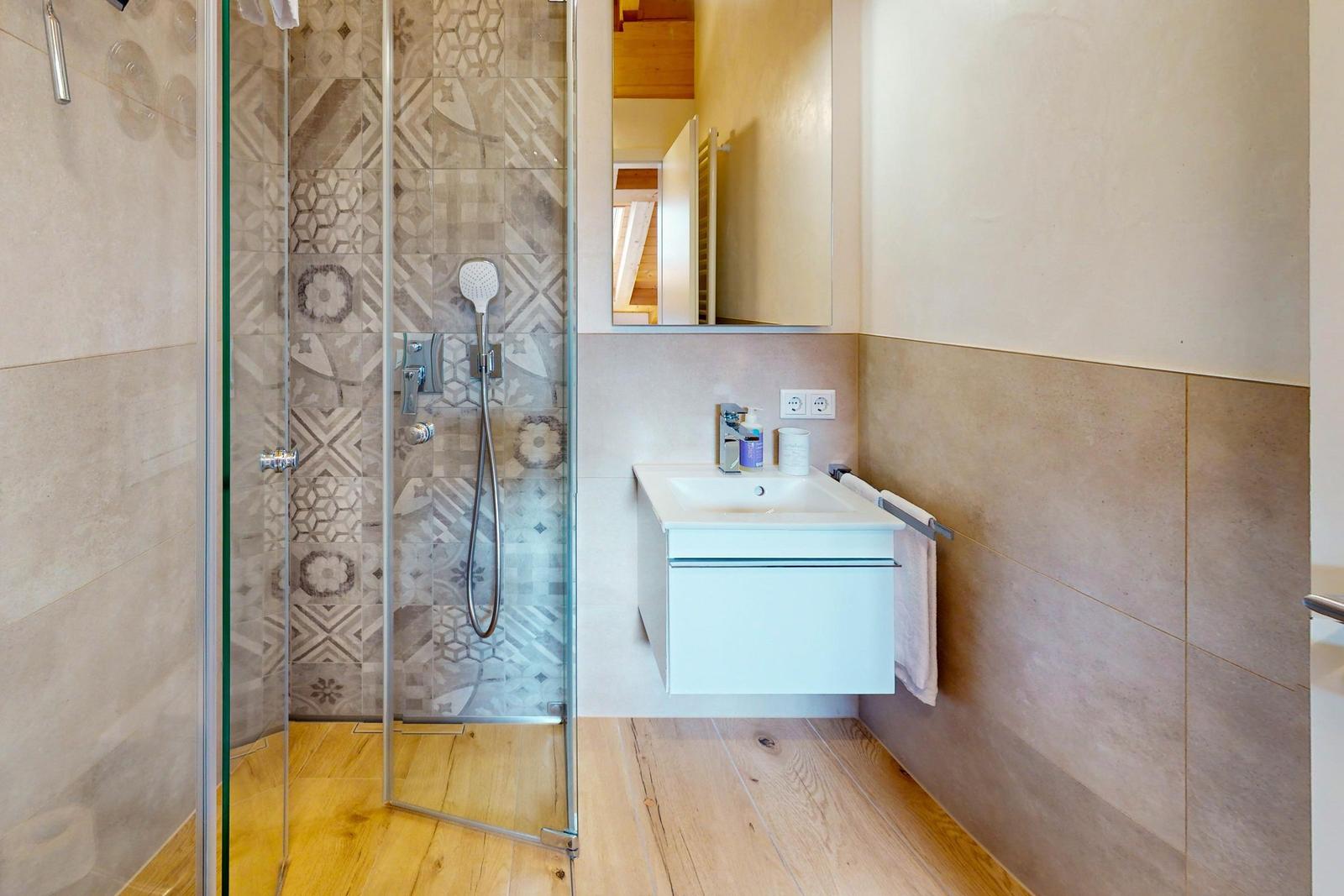 Dusche mit Glaswand, Waschbecken und Spiegel. Holzfußboden und Wandfliesen.