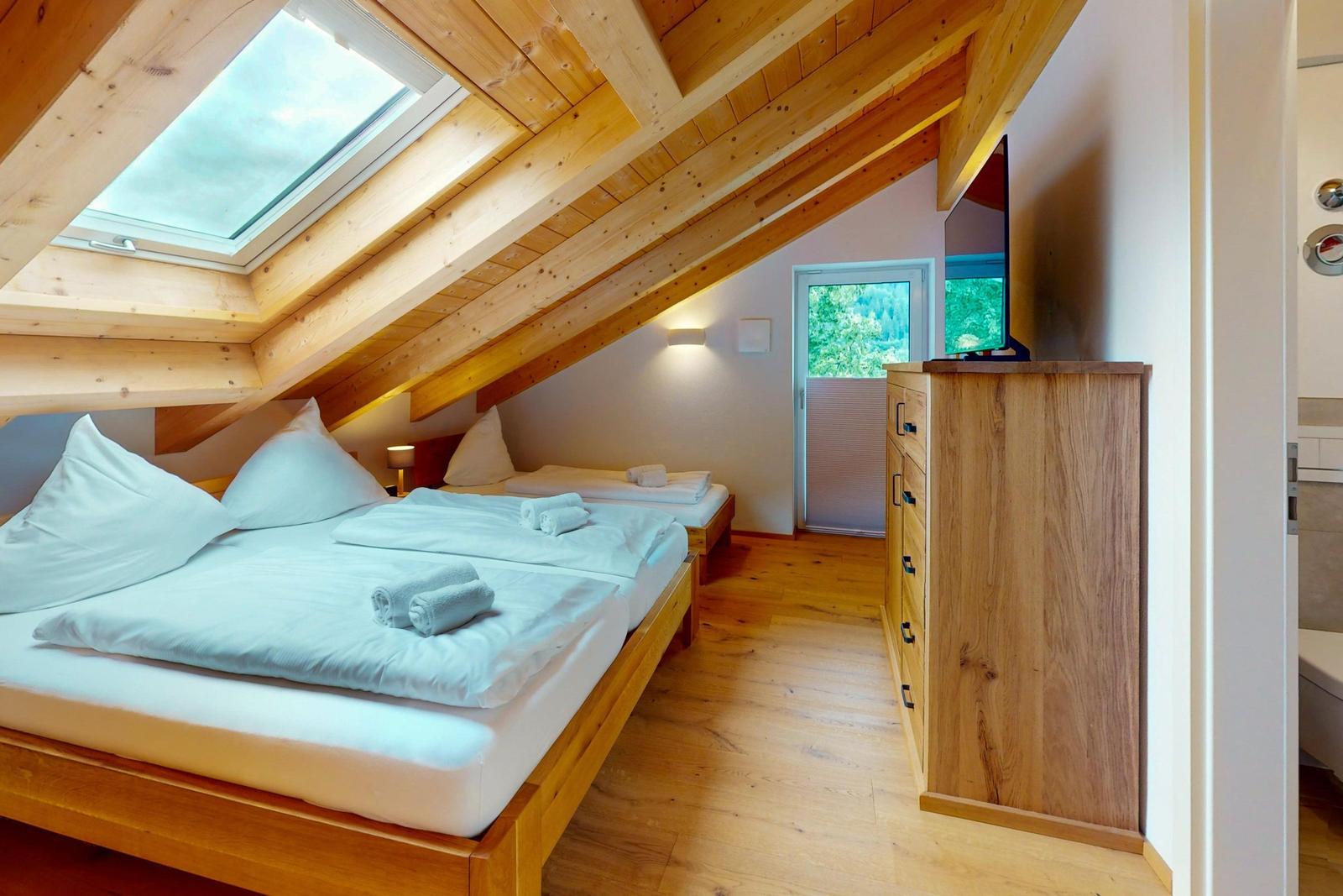 Schlafzimmer mit Doppelbett, kleinem Bett und Dachschrägen. Holzdecke und Fenster.