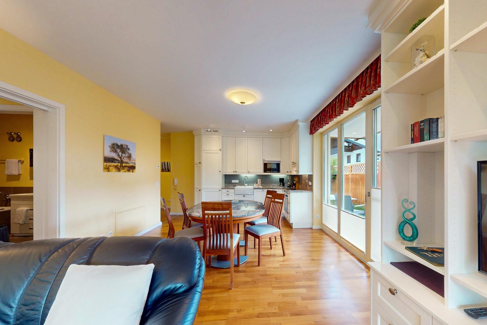 Wohnzimmer mit Essbereich und Küche. Holzboden, Sofa, Tisch mit Stühlen, Bücherregal und Fenster zur Terrasse.