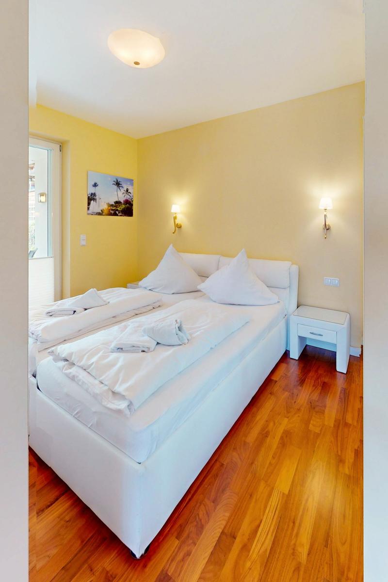 Schlafzimmer mit weißem Bett, Holzboden und gelben Wänden. An der Wand hängt ein Bild.
