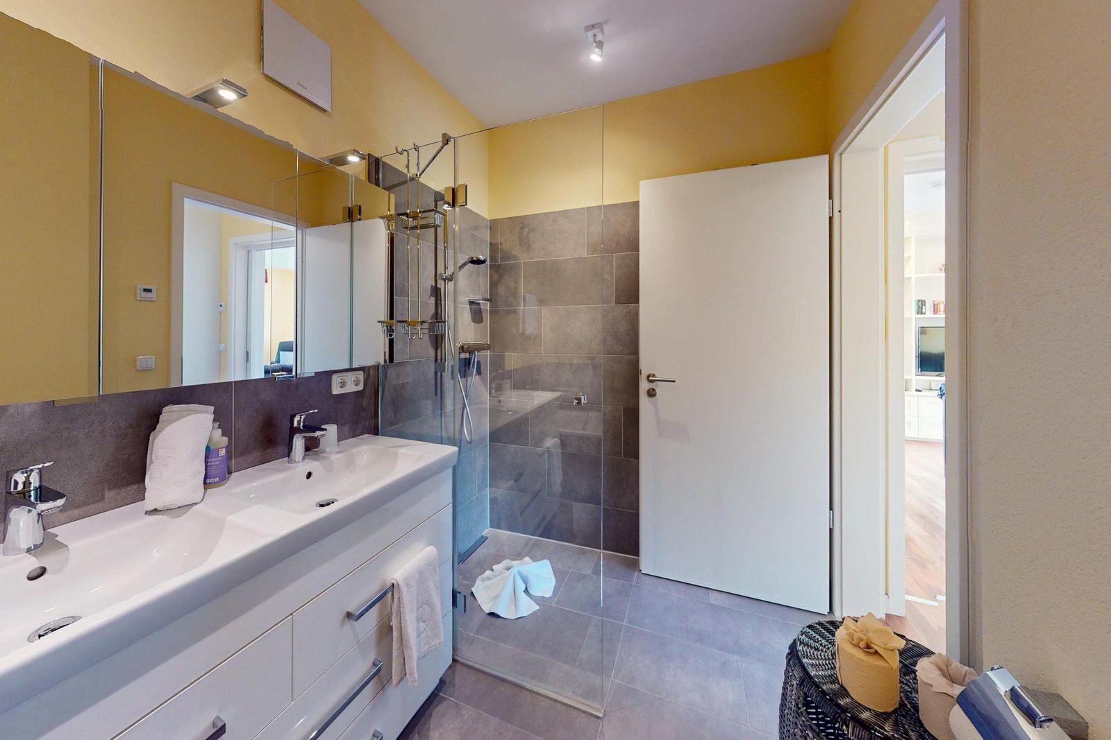 Badezimmer mit Dusche, Waschbecken und Spiegelschrank. Tür führt in Wohnbereich.