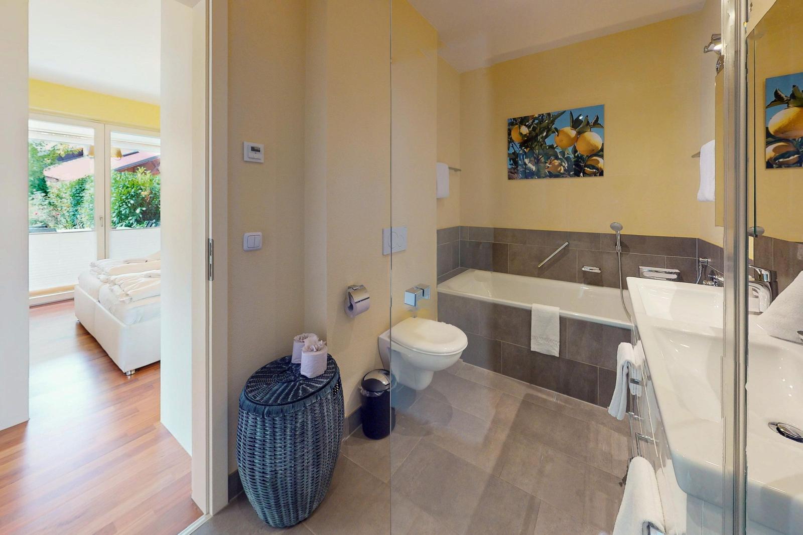 Badezimmer mit Badewanne, Waschbecken und Toilette. Durchgang zur Schlafzimmer mit Bett.