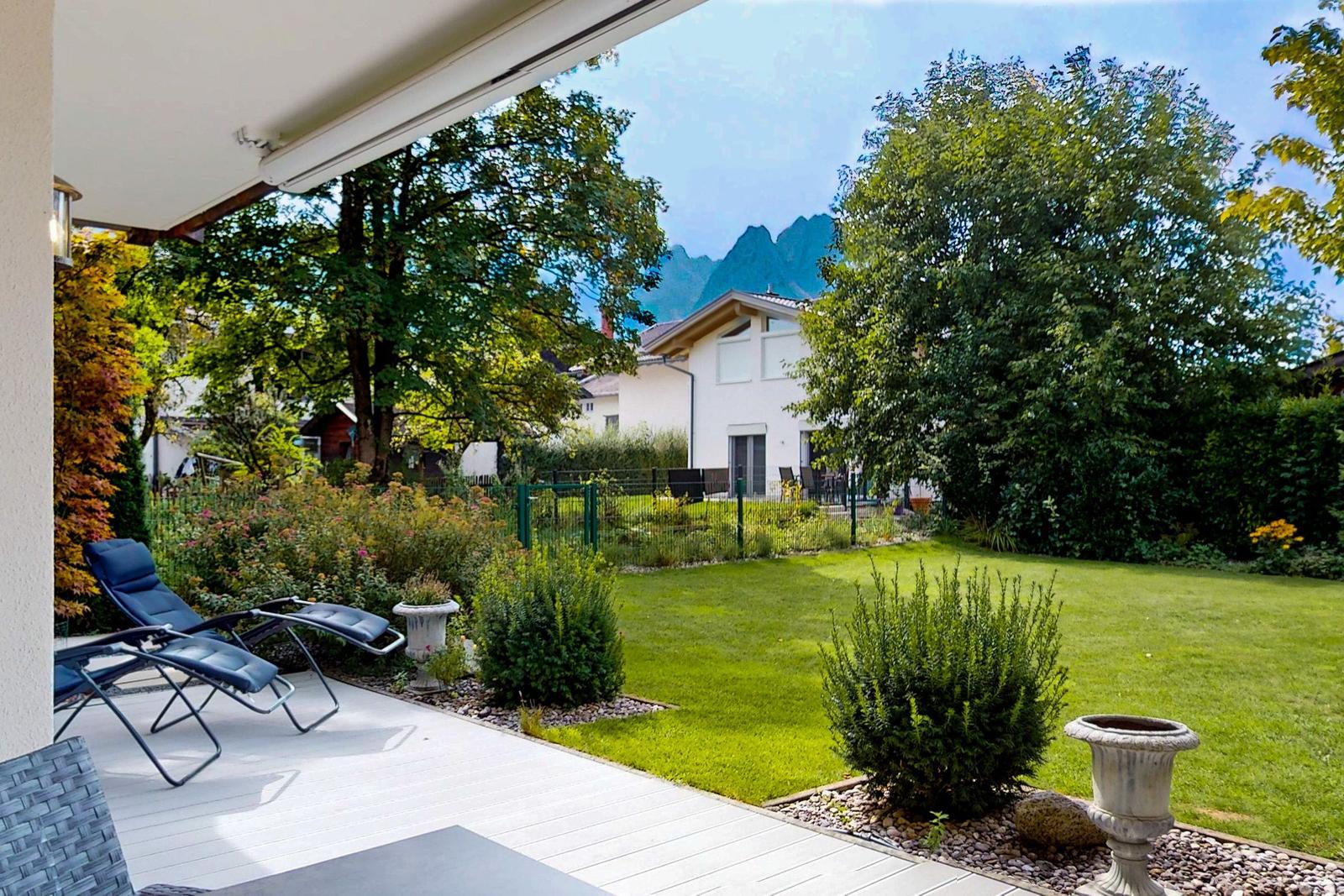 Terrasse mit Liegestühlen, Garten mit Rasen und Bäumen, Bergpanorama im Hintergrund.