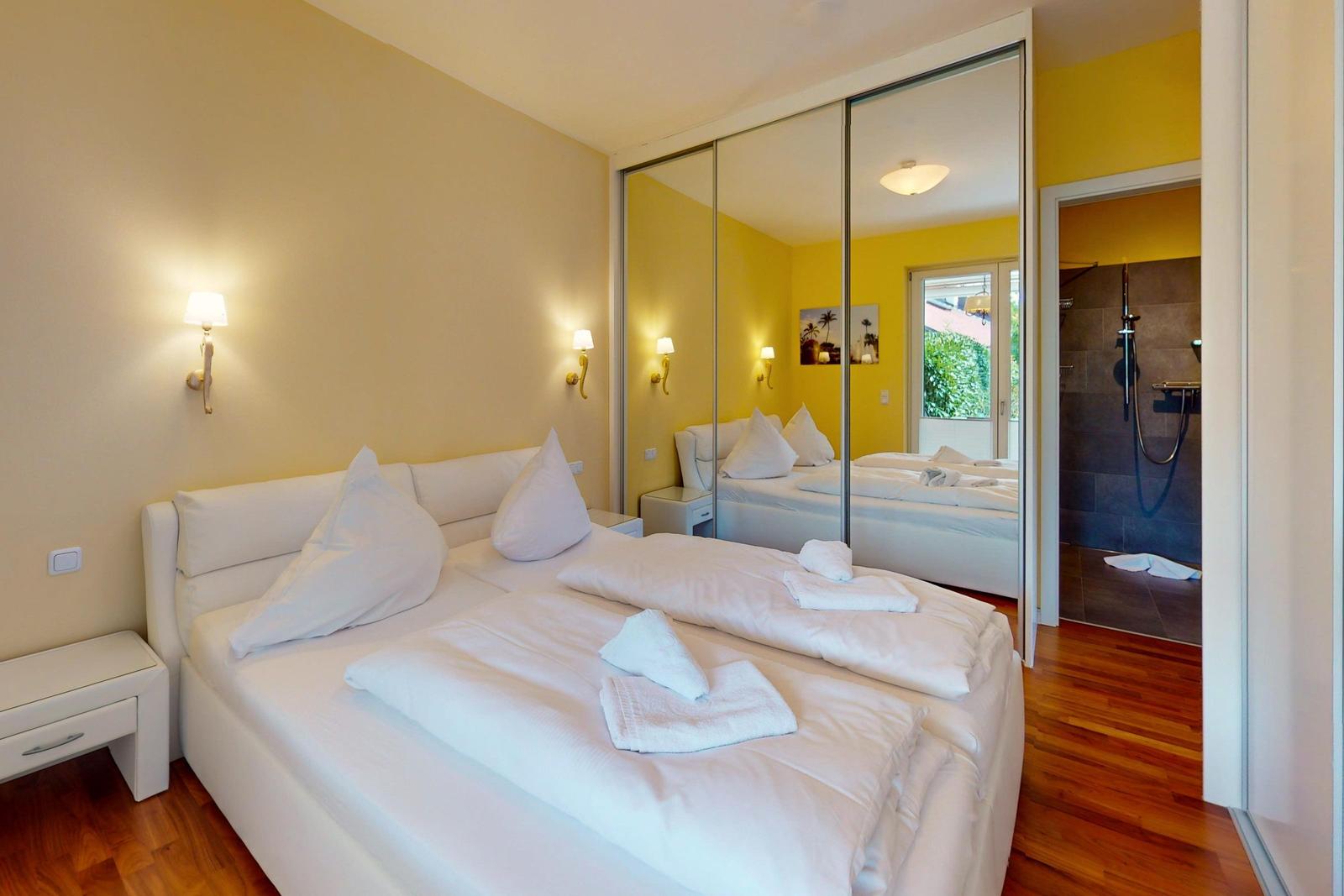 Schlafzimmer mit weißem Bett, Spiegelschrank und angrenzender Dusche.