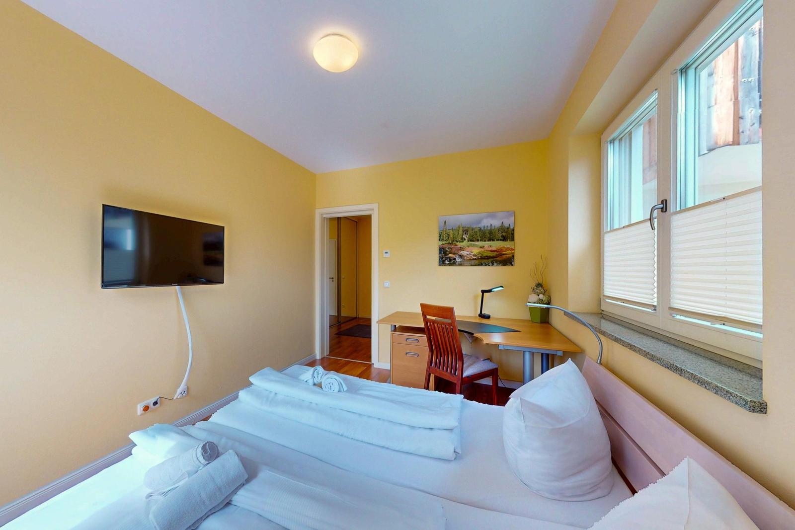 Zimmer mit Bett, Schreibtisch, Fernseher und Fenster mit Jalousien.