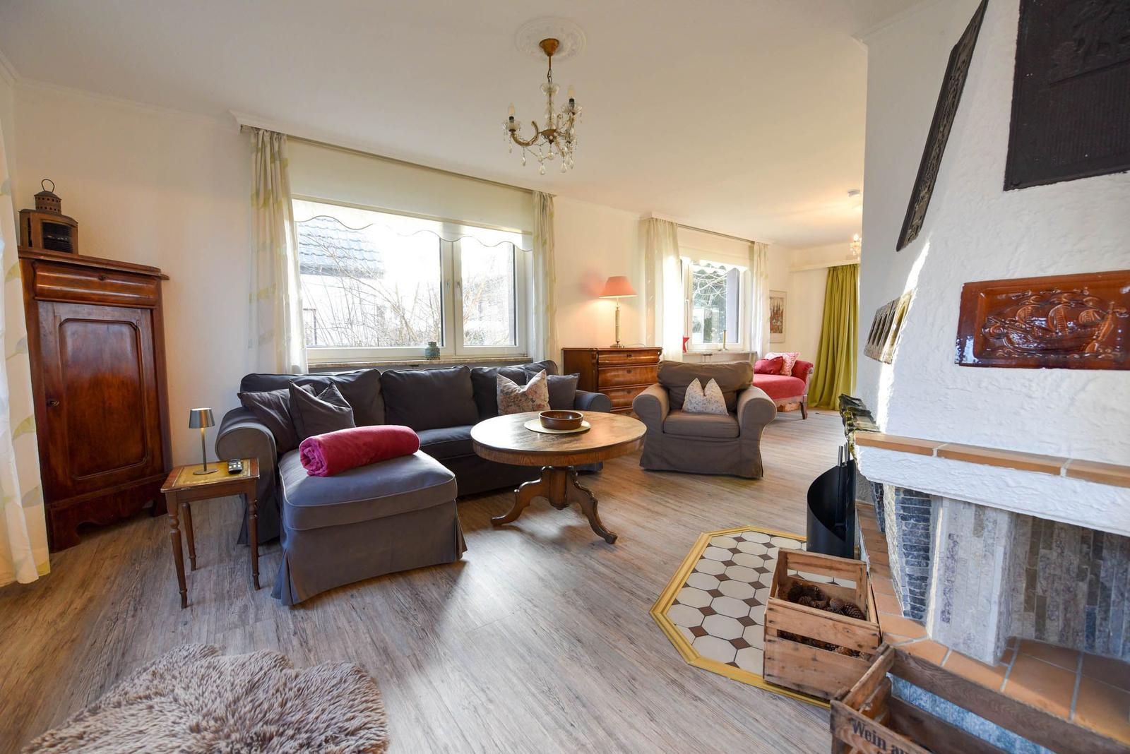 Wohnzimmer mit Sofa, Sessel, Kamin und Holzboden. Fenster mit Vorhängen und Kronleuchter.