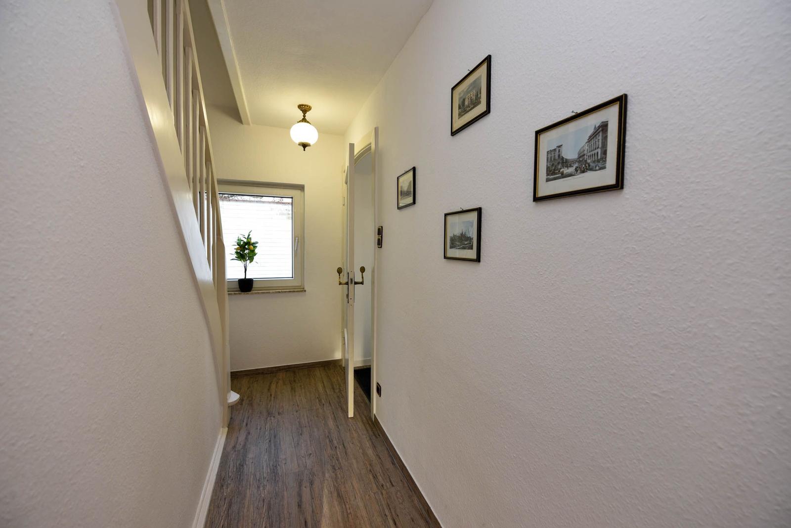 Flur mit Holzboden, Wandbildern und Treppenhaus. Fenster mit Pflanze.