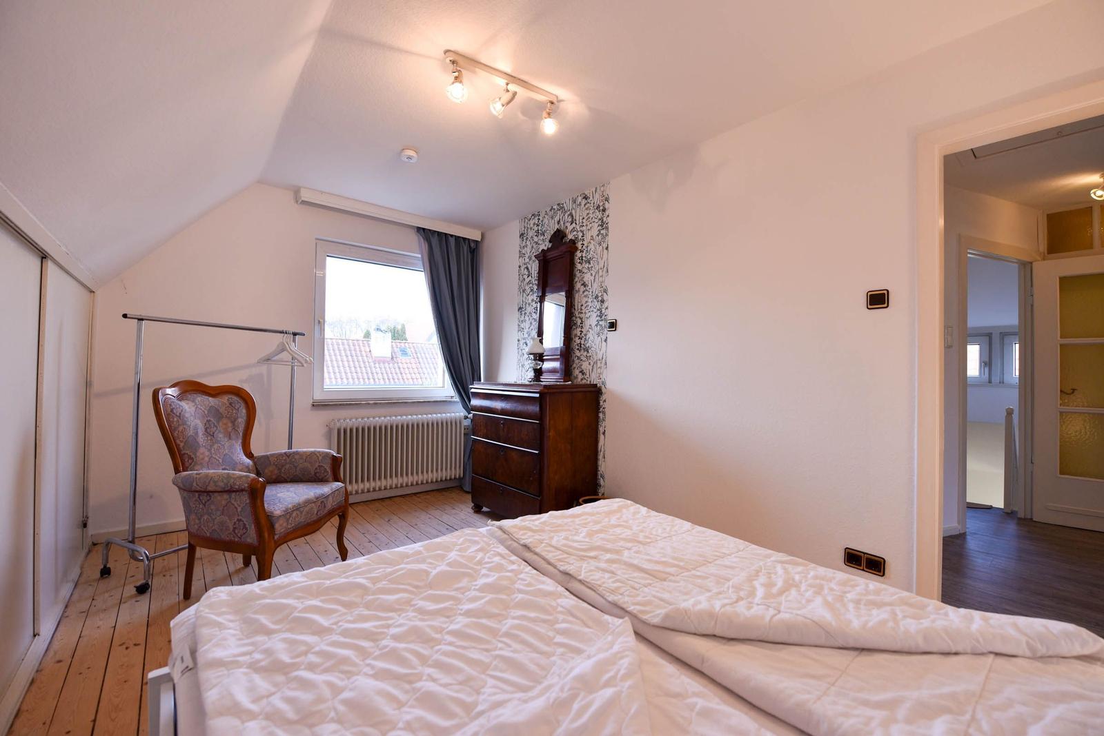 Schlafzimmer mit Doppelbett, Stuhl, Schrank und Fenster mit Blick auf Dach.