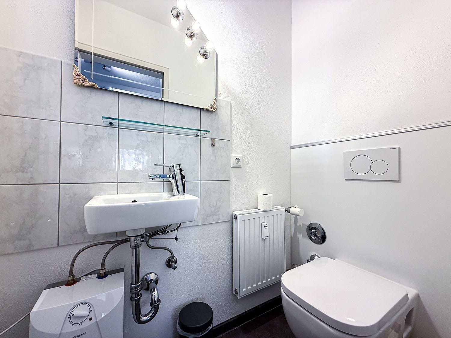 Badezimmer mit Waschbecken, Spiegel und Toilette. Wandheizung und Heizungsgerät sichtbar.