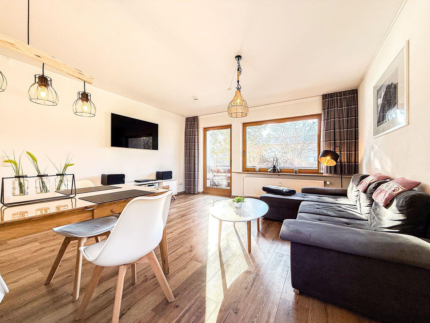 Wohn- und Essbereich mit Sofa, Tisch und TV. Holzfußboden und große Fenster.