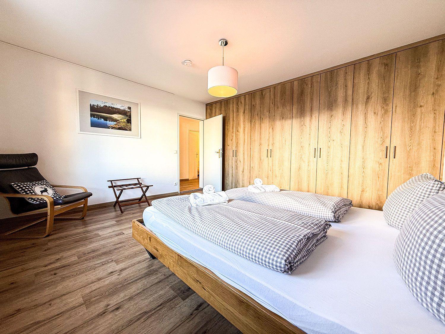 Schlafzimmer mit Doppelbett, Holzwand und Sessel. Auf dem Bett liegen Handtücher.