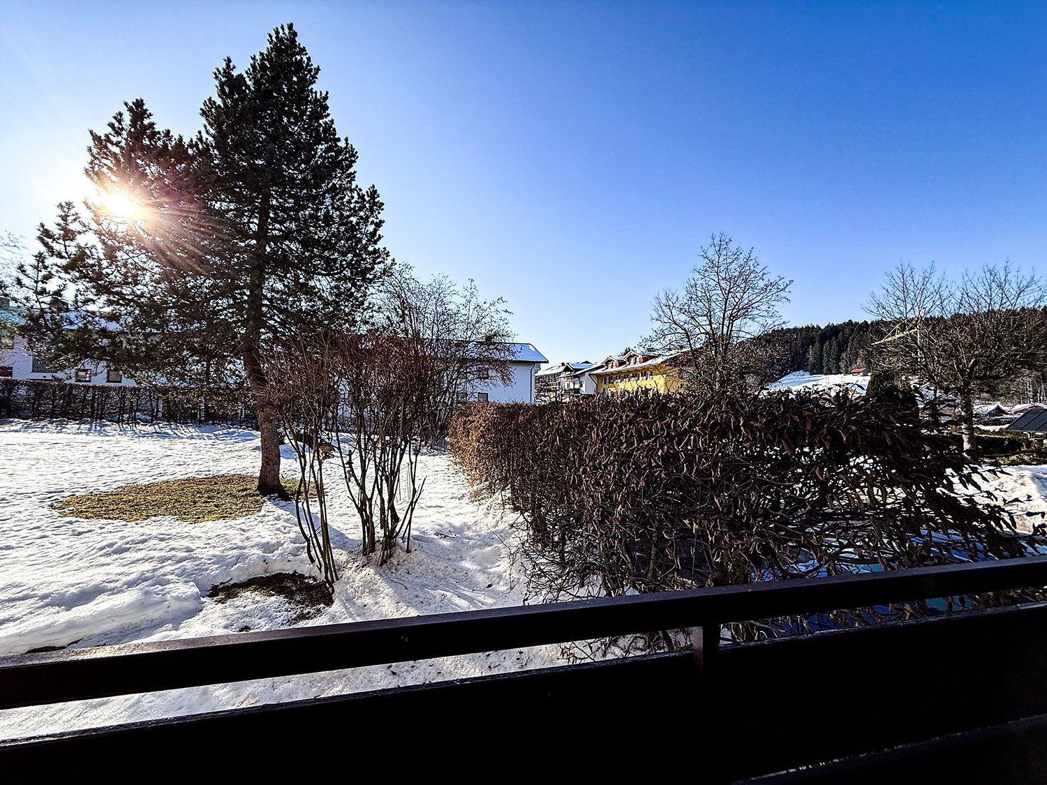 Schneebedeckter Garten mit Baum und Hecke, Blick auf Häuser und Berge unter blauem Himmel.