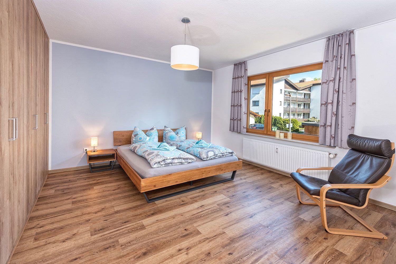 Schlafzimmer mit Doppelbett, Holzboden und Sessel. Fenster mit Vorhängen und Blick auf Gebäude.