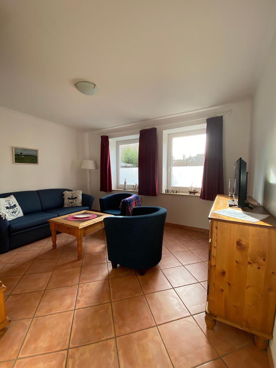 Wohnzimmer mit Sofa, zwei Sesseln, Holztisch und TV-Schrank. Fenster mit roten Vorhängen.