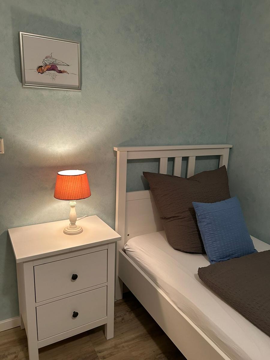Schlafzimmer mit weißem Bett, Nachttisch und Lampe. Wandbild über dem Bett.