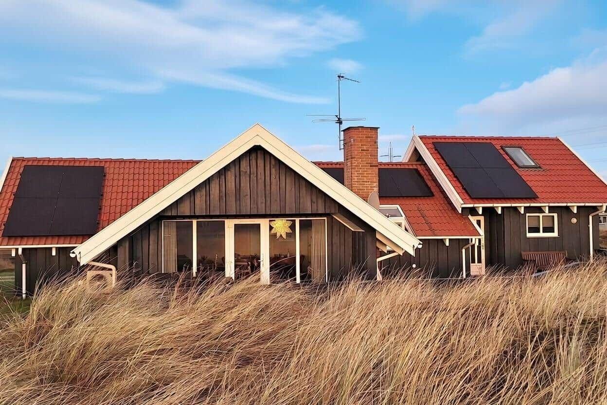 Holzhaus mit rotem Dach und Solarpanelen im Hintergrund. Vorne hohes Gras.