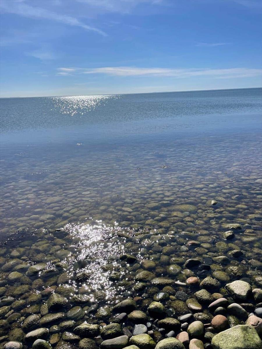 Klarer See mit Steinen im Vordergrund und Sonnenreflexionen auf der Wasseroberfläche.
