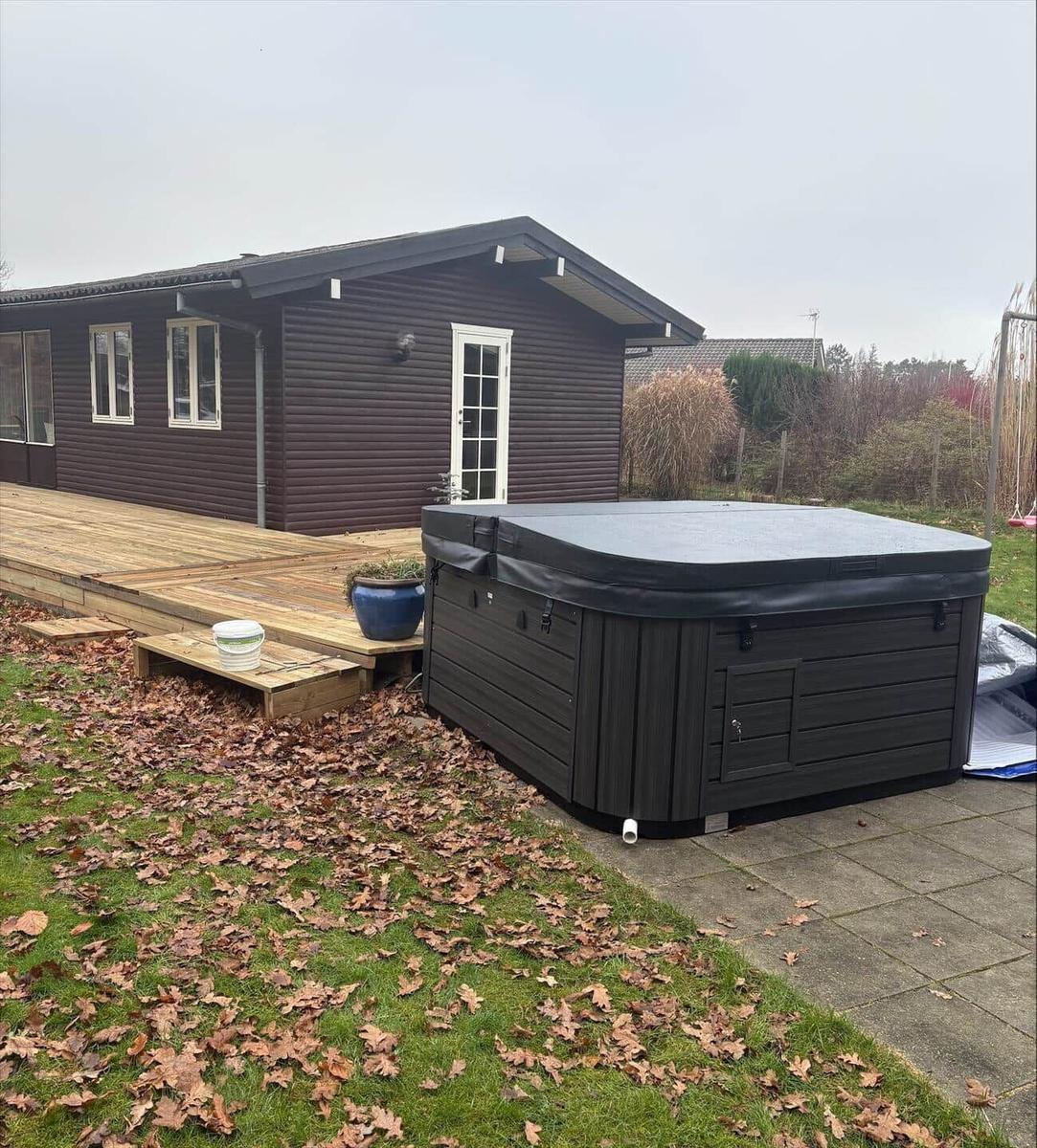 Schwarzer Whirlpool auf Terrasse neben Holzhaus mit grünem Rasen und herbstlichen Blättern.