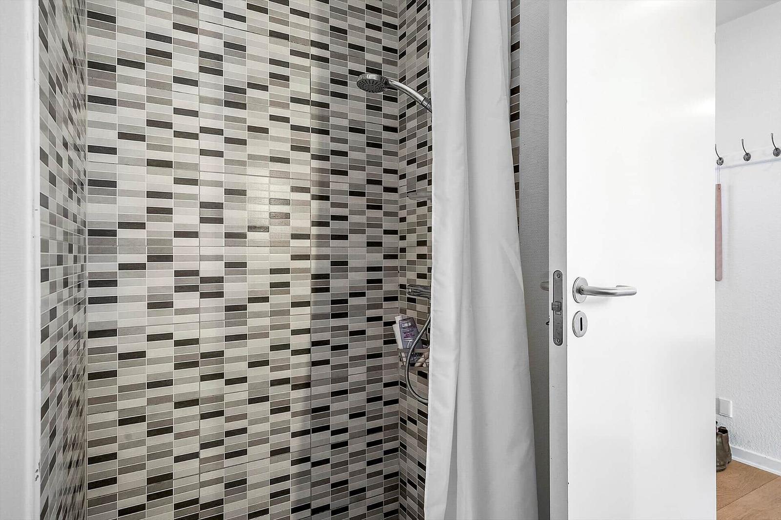 Dusche mit Mosaikfliesen und Duschvorhang. Tür und Wand mit Haken sichtbar.