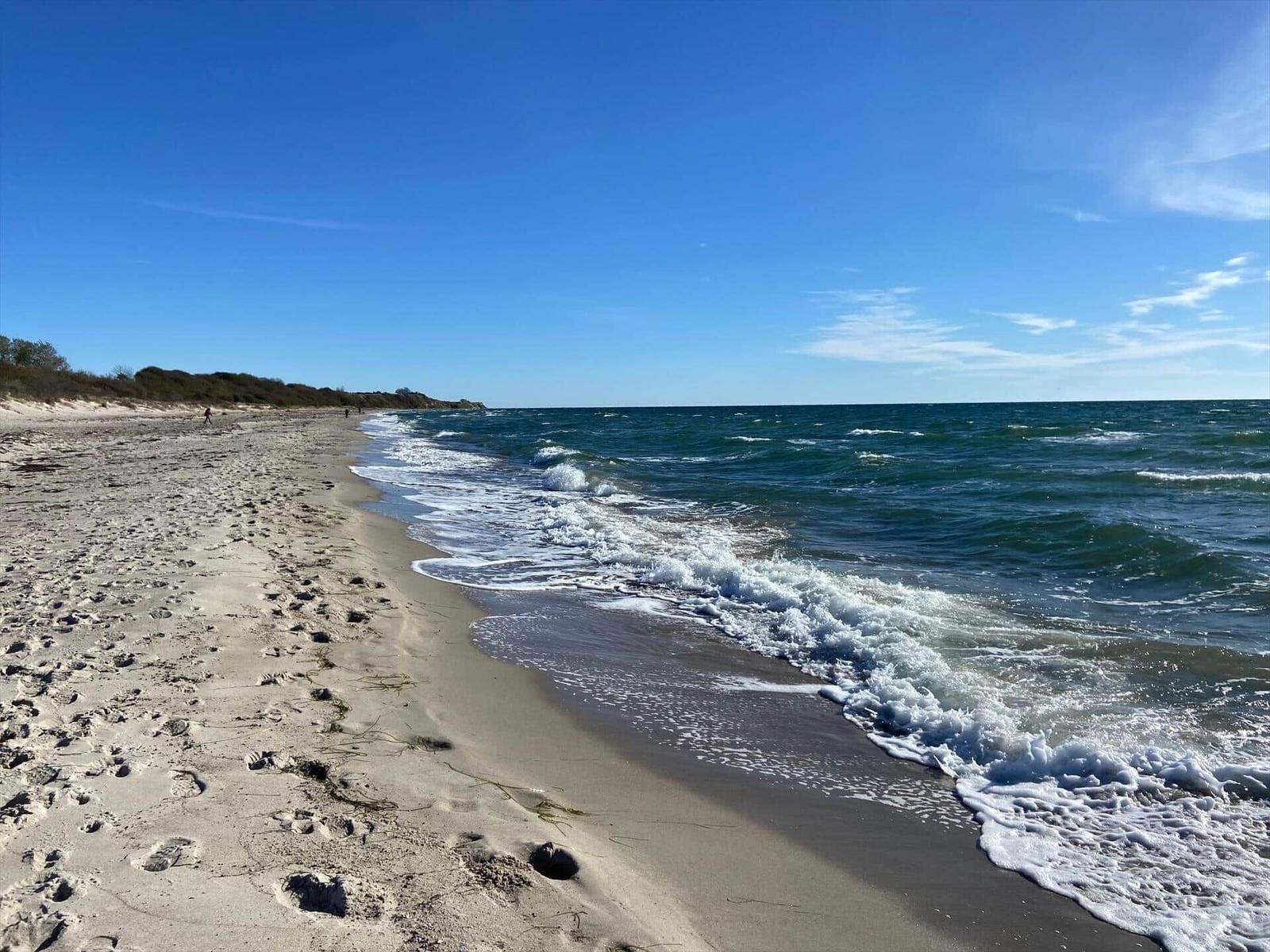 Strand mit Wellen, Sand und klarem Himmel. Ein paar Menschen sind fern sichtbar.