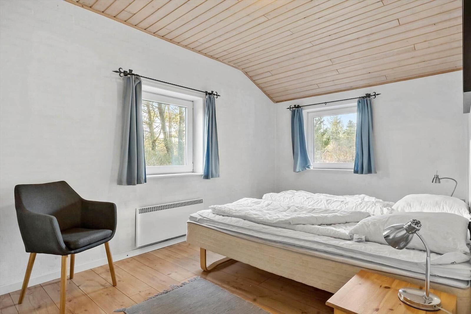 Schlafzimmer mit Bett, Stuhl und zwei Fenstern mit Vorhängen.