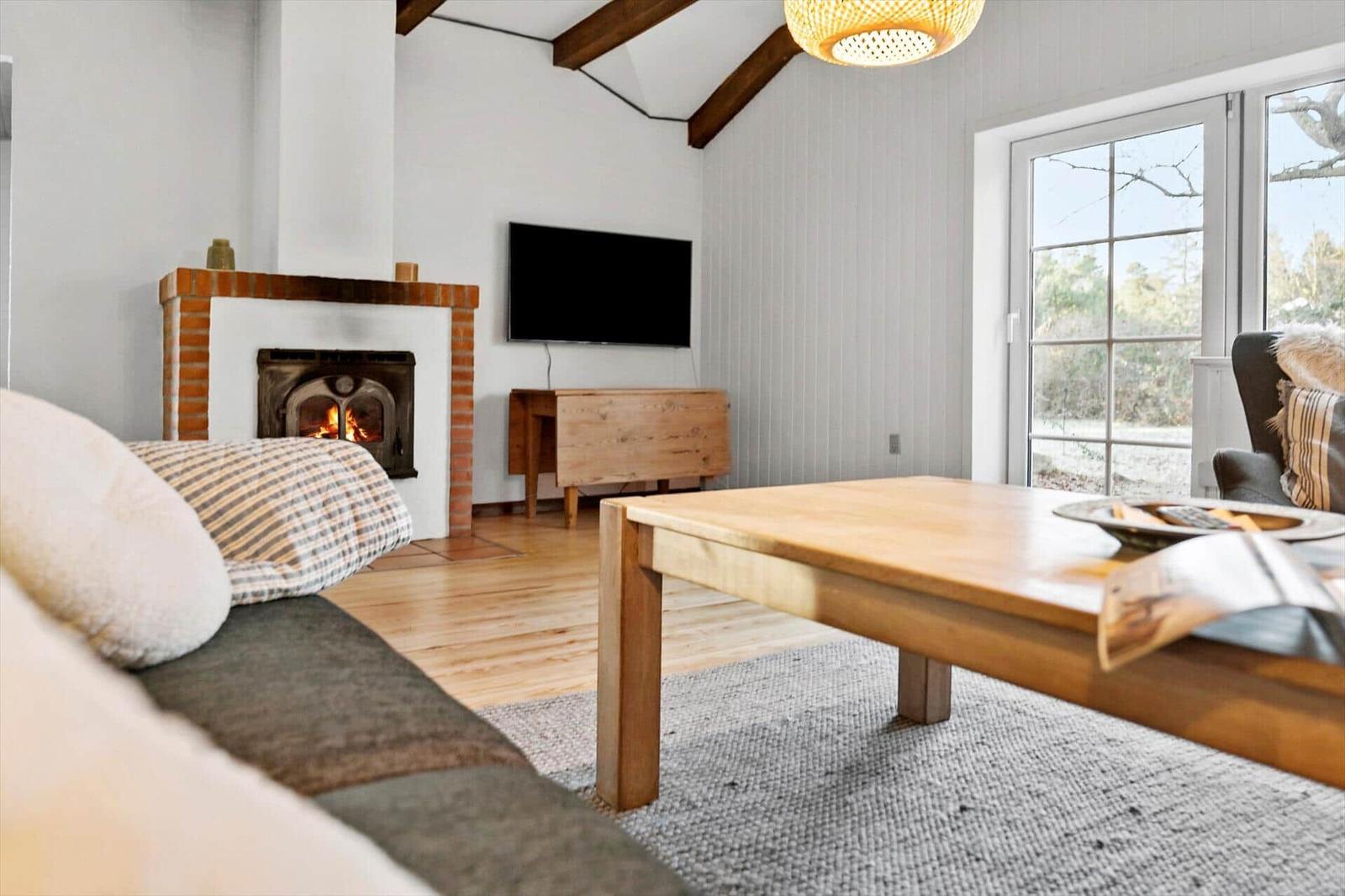 Wohnzimmer mit Kamin, TV und Holztisch. Große Fenster mit Blick auf Außenbereich.