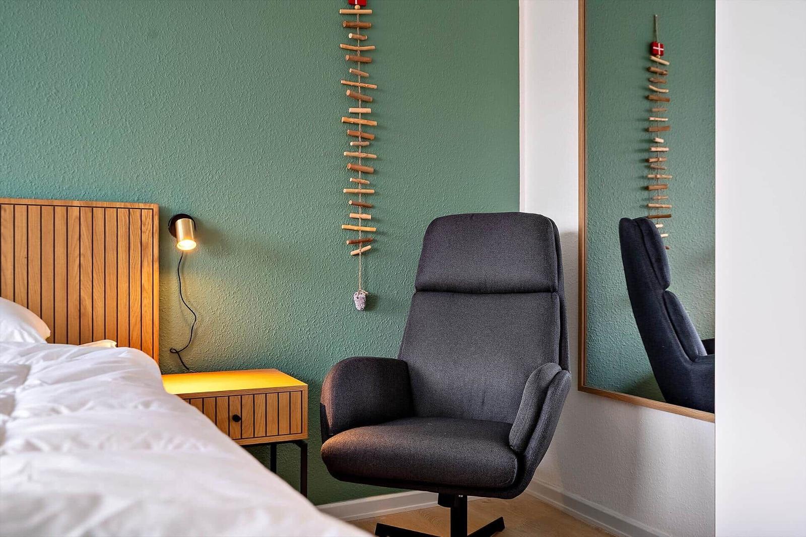 Schlafzimmer mit Bett, Nachttisch und Sessel. Wanddekoration und Spiegel sichtbar.