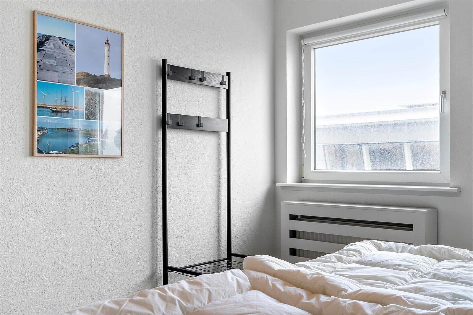 Schlafzimmer mit Bett, Kleiderständer und Fenster mit Blick auf eine Mauer.