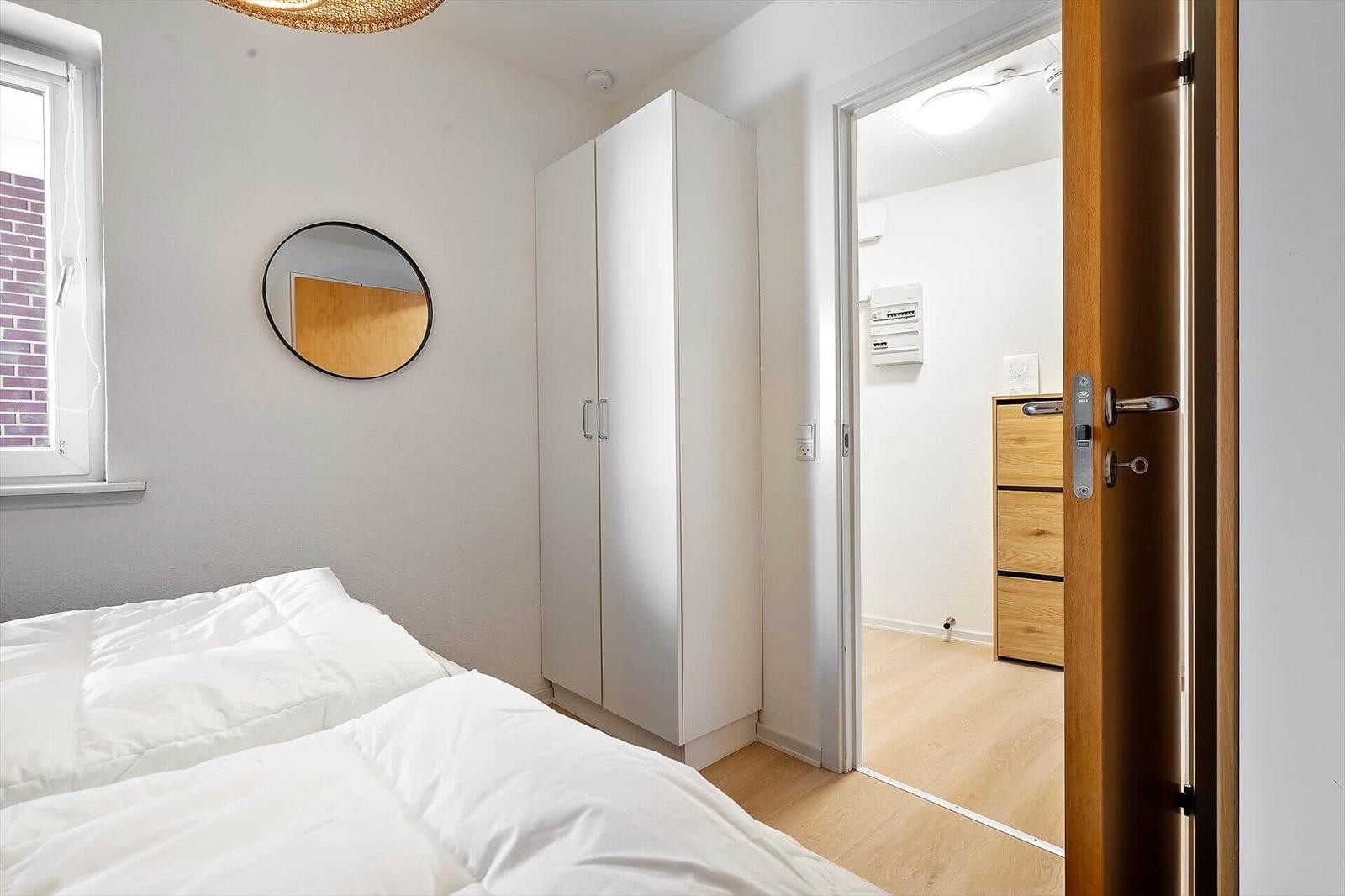 Schlafzimmer mit weißem Bett, Kleiderschrank und runden Spiegel. Durchgang zu angrenzender Raum.