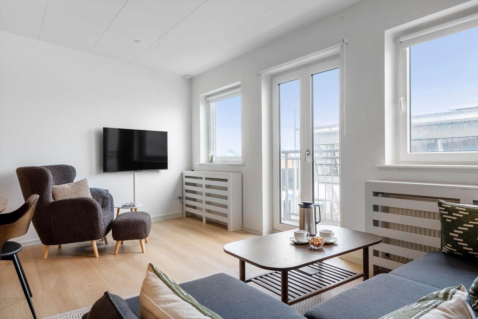 Wohnzimmer mit Sofa, Sessel, Couchtisch und Fernseher. Große Fenster und Balkontür.