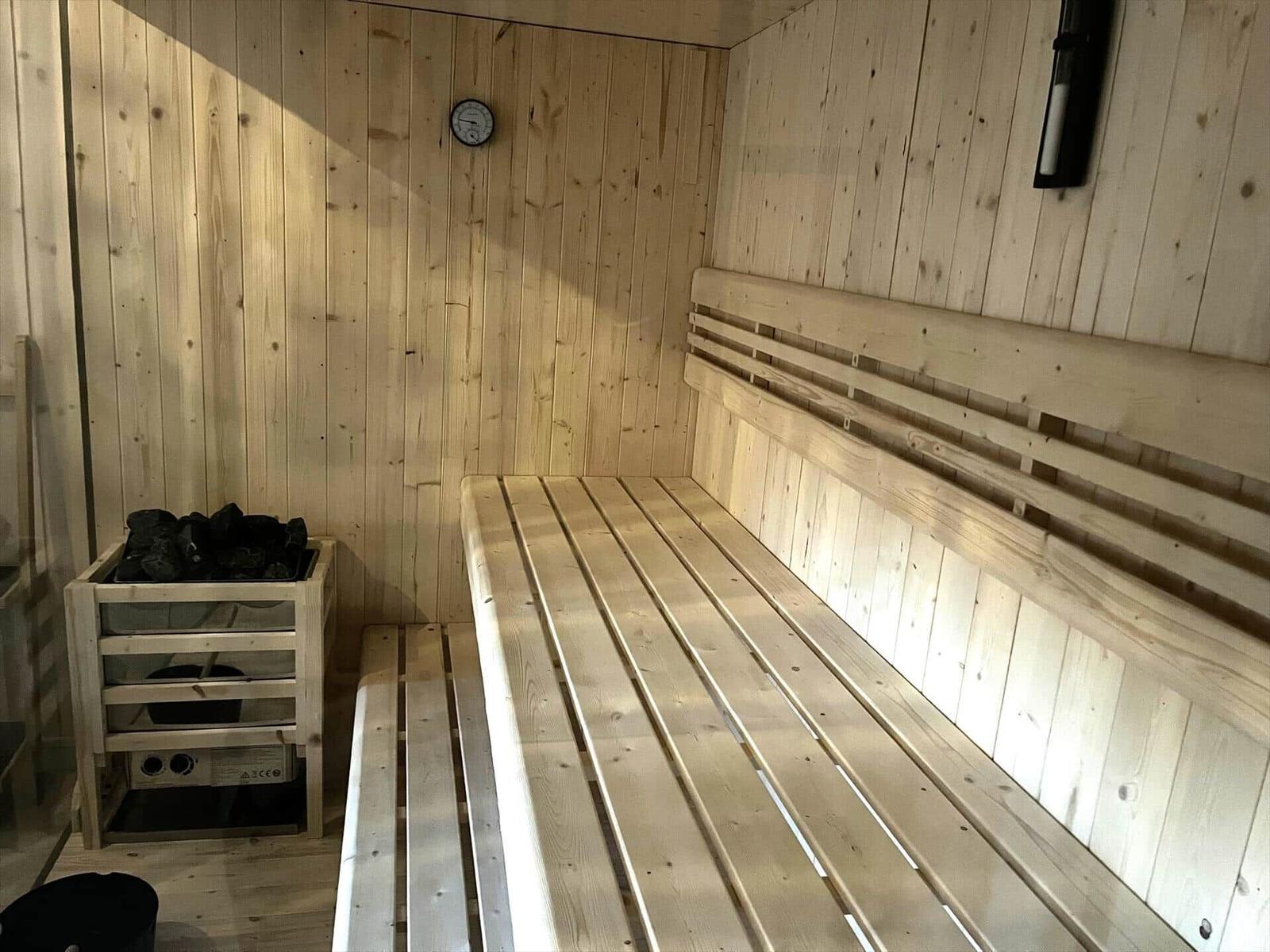 Sauna with wooden bench and stove, wall clock and light switch visible.