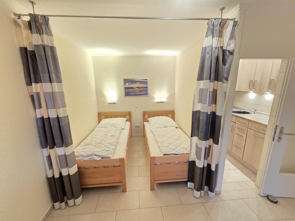 Zwei Einzelbetten mit Holzrahmen und gestreiften Vorhängen. Küche im Hintergrund sichtbar.