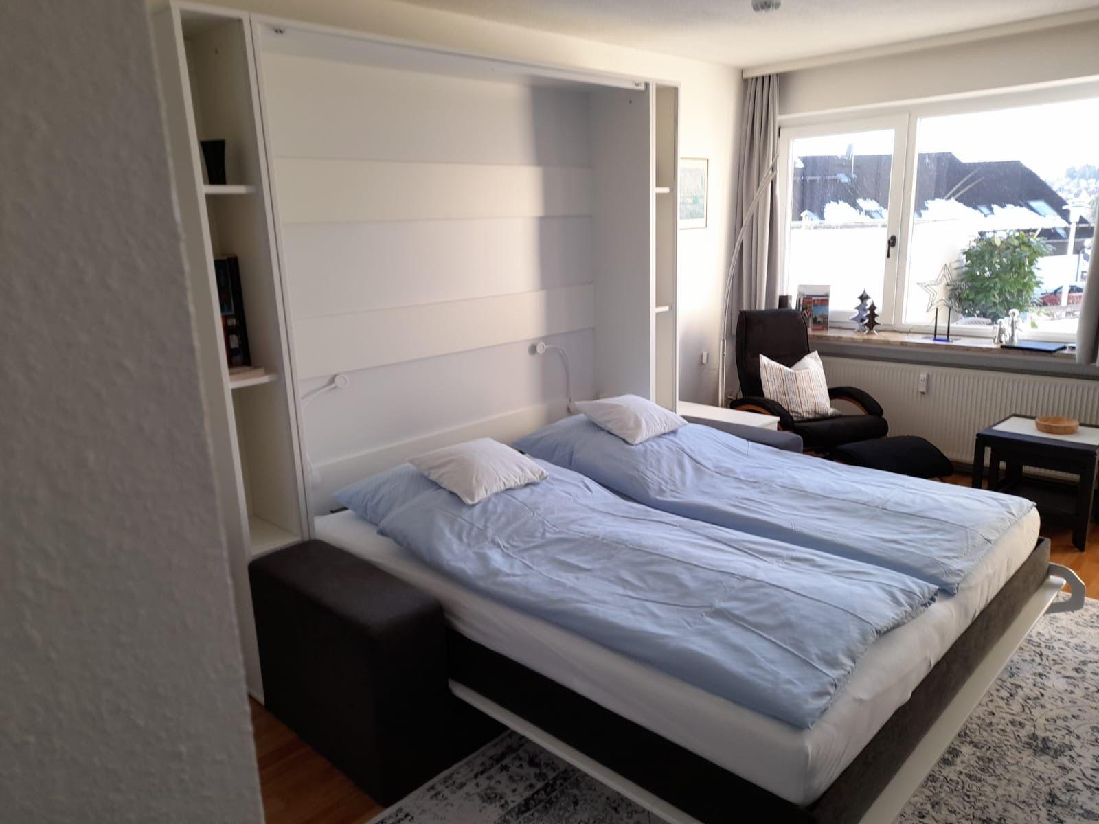 Schlafzimmer mit Bett, Sessel und Fenster. Wandbett und Regal sichtbar.