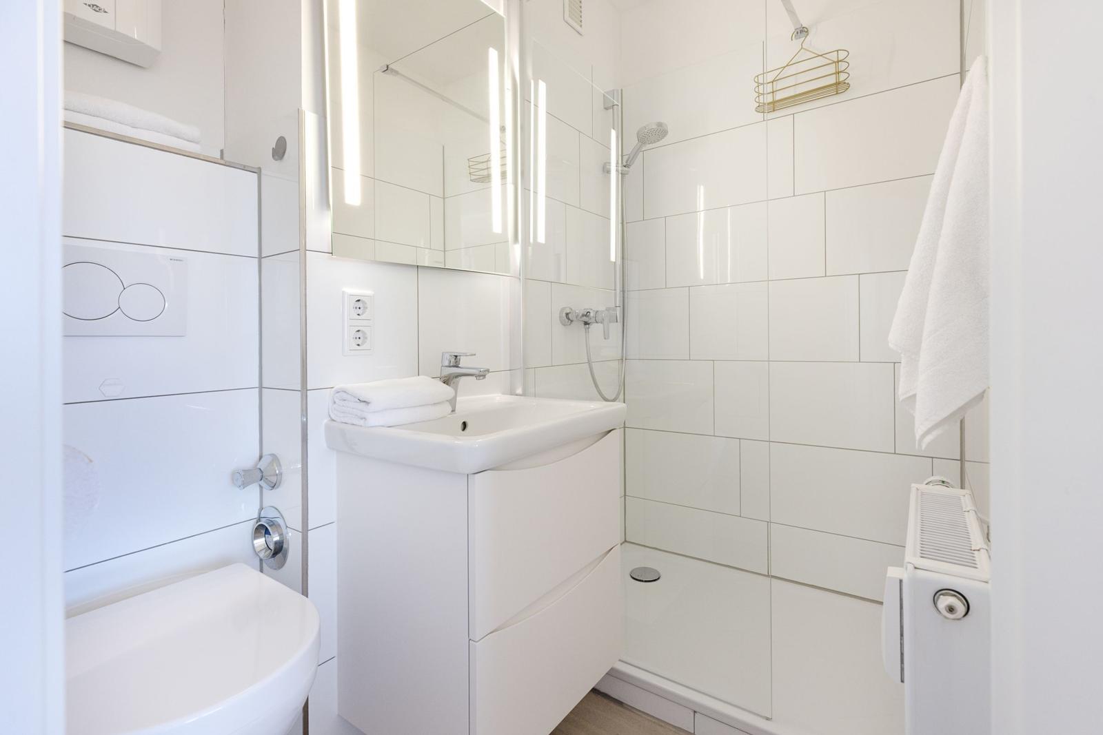 Badezimmer mit Dusche, Waschbecken und Toilette. Weiße Fliesen und moderne Einrichtung.