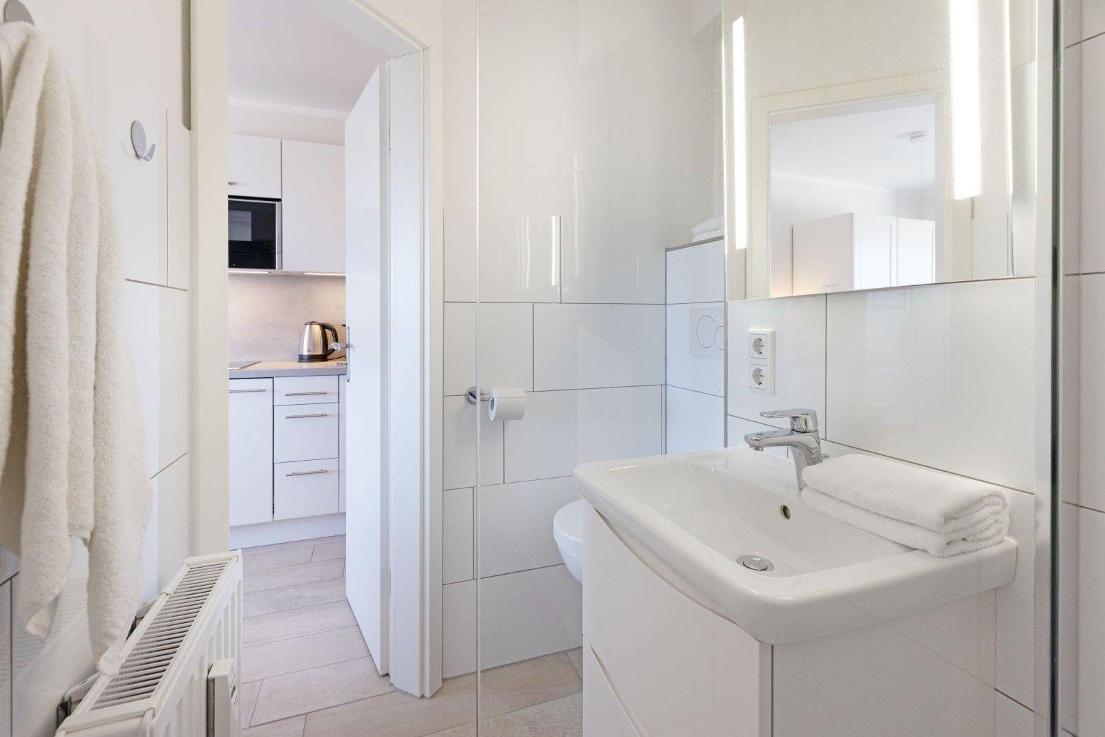 Badezimmer mit Waschbecken, Spiegel und Toilette. Küche sichtbar im Hintergrund.