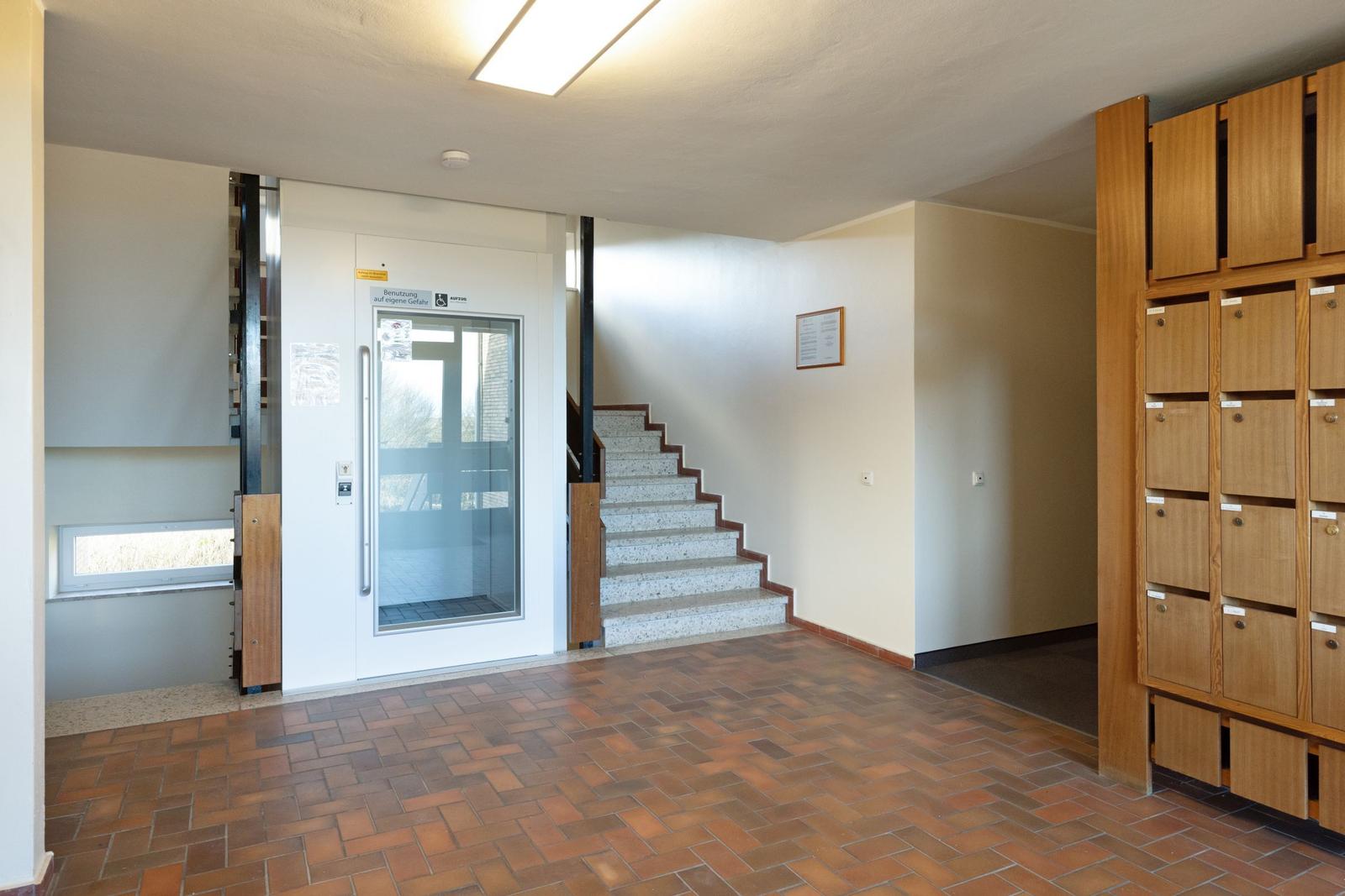 Eingangsbereich mit Aufzug, Treppe und Holzbriefkastenwand.