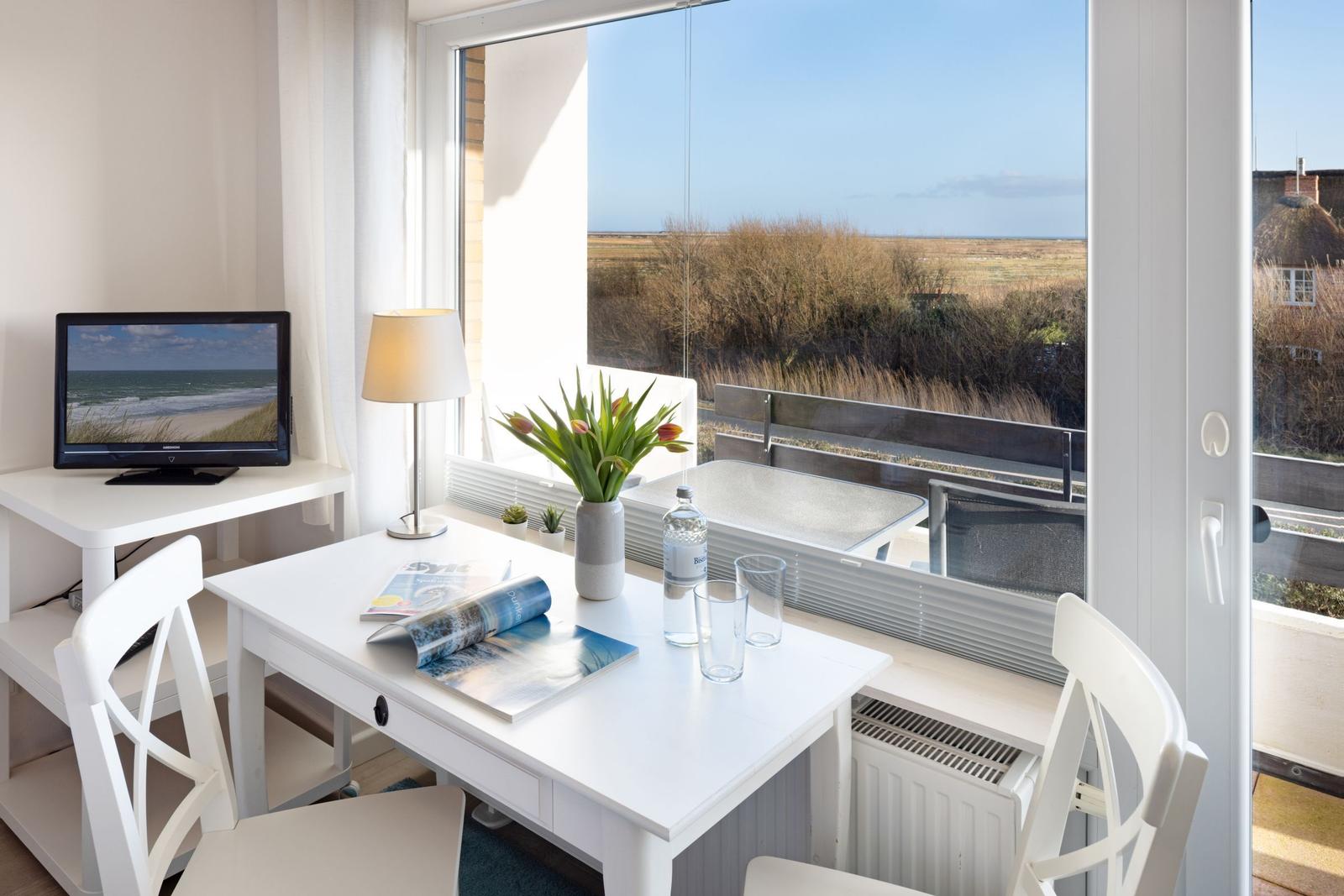 Wohnraum mit Tisch, Stühlen und TV. Durch Fenster Blick auf Strand und Dünen.