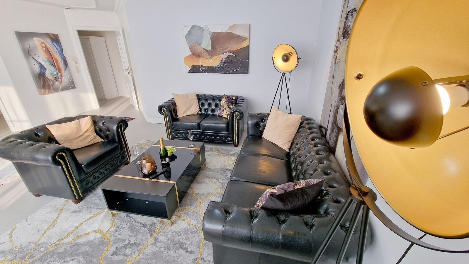 Wohnzimmer mit schwarzen Ledersofas, goldfarbenen Lampen und abstrakten Wandbildern.