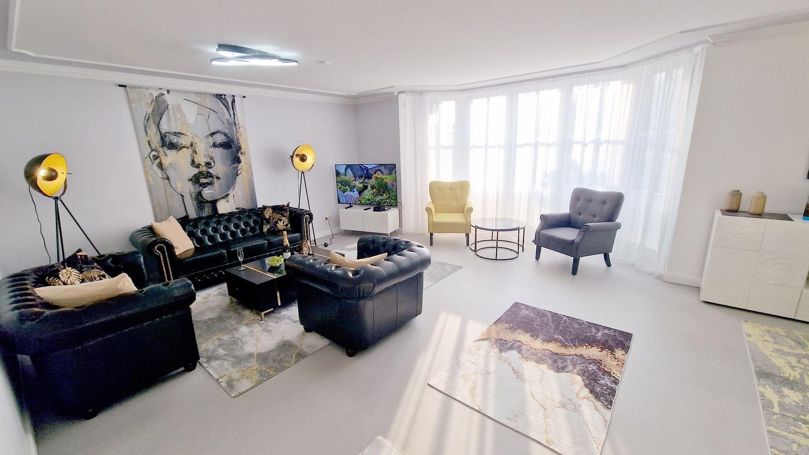 Wohnzimmer mit Ledersofa, zwei Sesseln und großem Fenster. Wandbild und Teppiche sorgen für Stil.