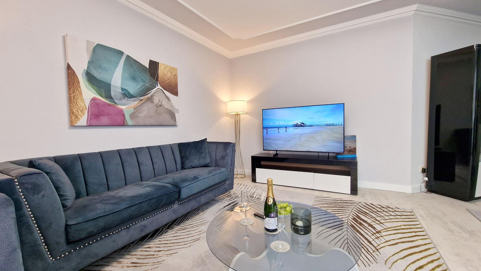 Ein Wohnzimmer mit blauem Sofa, Glas-Tisch und Fernseher auf schwarz-weißem Regal.