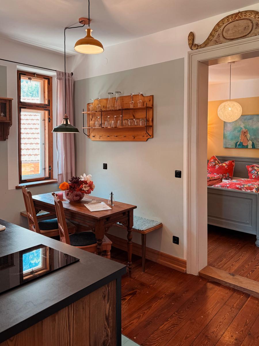 Essbereich mit Holztisch, Stühlen und Wandregal. Durchgang zu Schlafzimmer mit Bett und Wandbild.