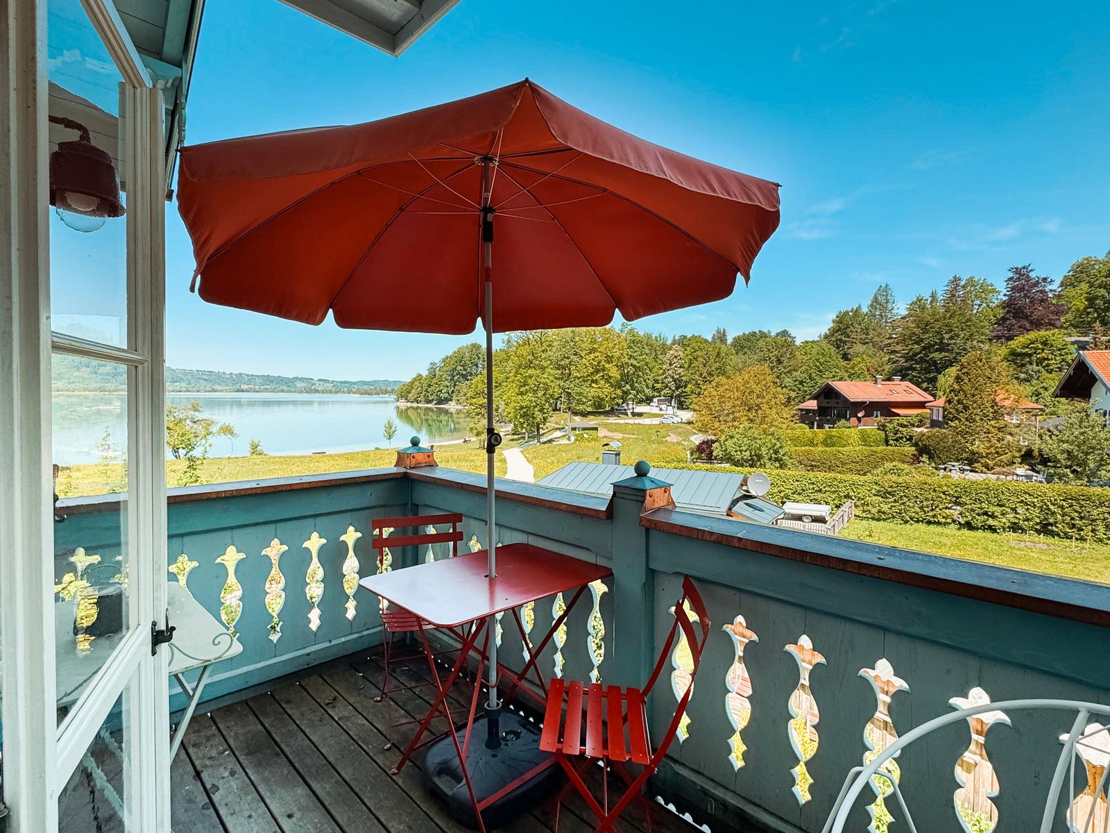 Terrasse mit rotem Sonnenschirm und Tisch. Blick auf See und grüne Landschaft.