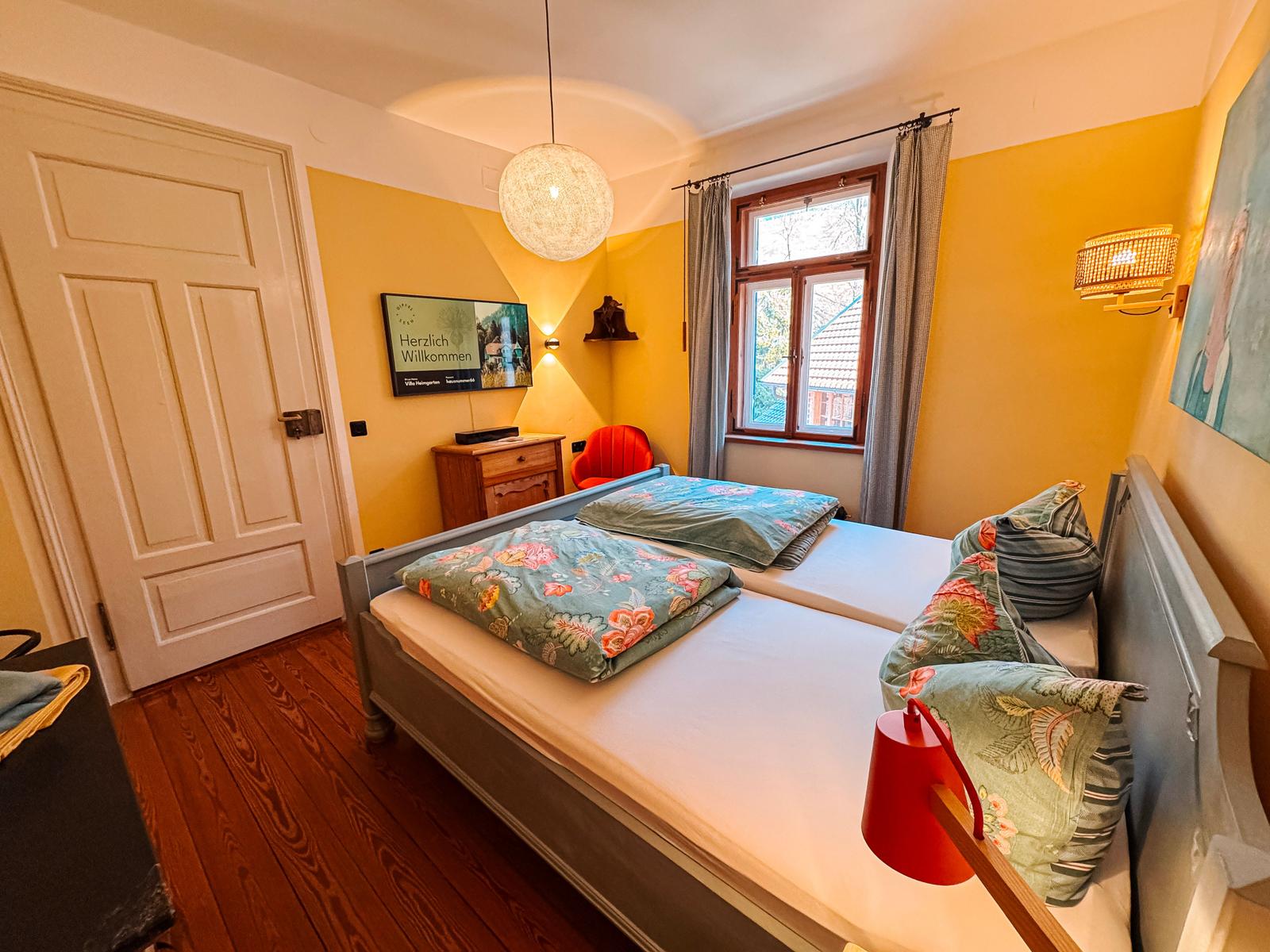 Schlafzimmer mit Doppelbett, Fenster, TV und Holzboden.