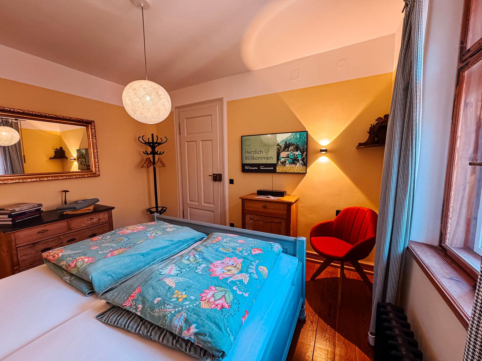 Schlafzimmer mit Bett, Fernseher, Spiegel und roter Sitzgelegenheit.