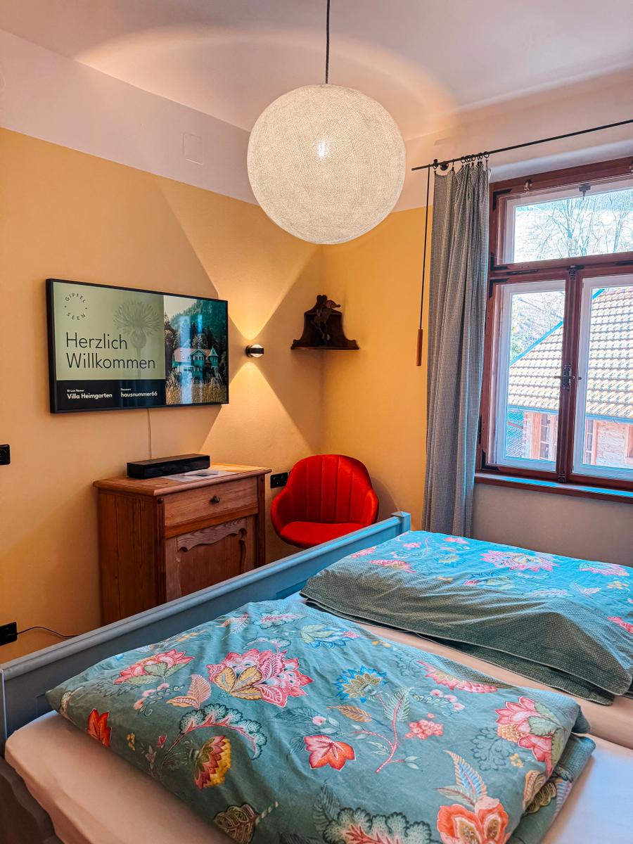 Schlafzimmer mit Bett, TV und Fenster. Blaue Bettwäsche mit Blumenmuster.