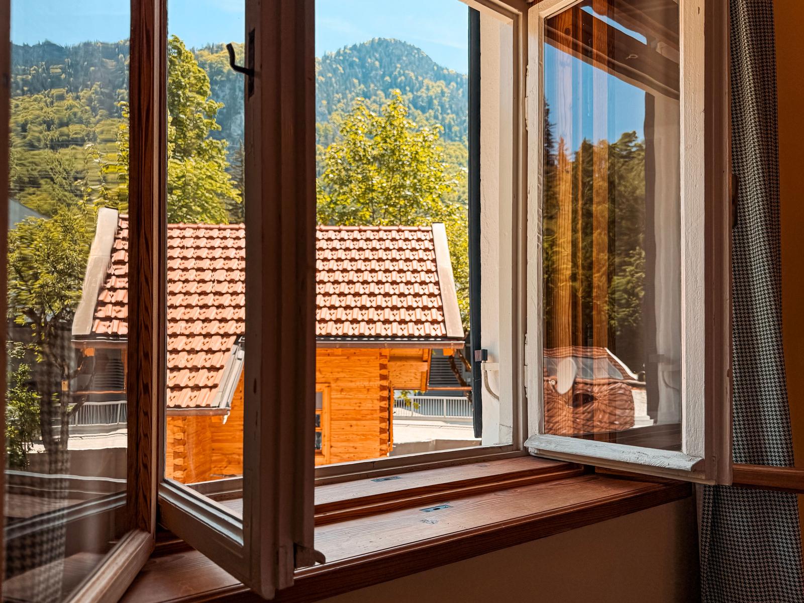Durch das Fenster blickt man auf ein Holzhaus mit rotem Dach und grüne Berghänge.