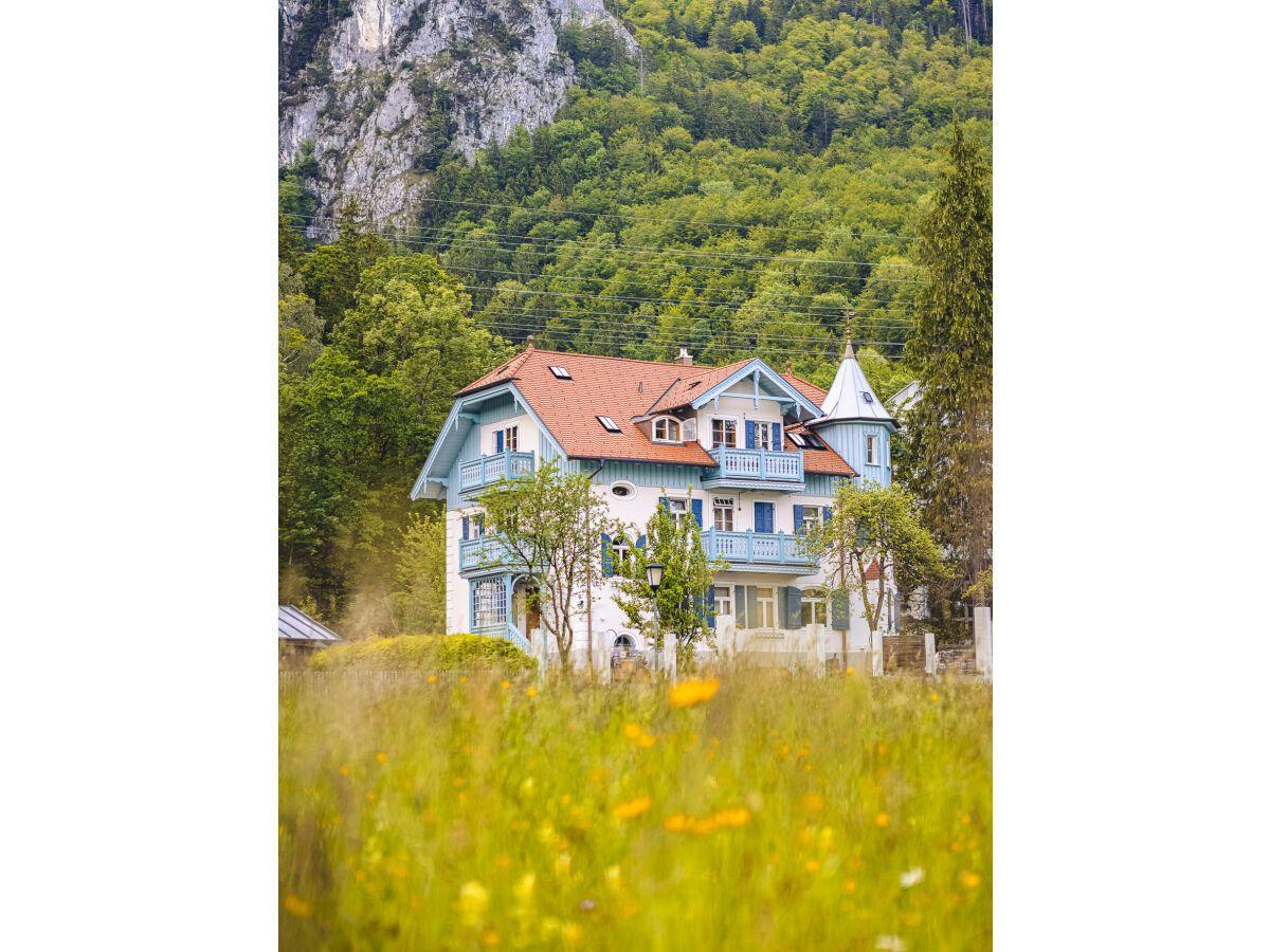 Haus mit rotem Dach und blauen Balkonen steht in grüner Umgebung.
