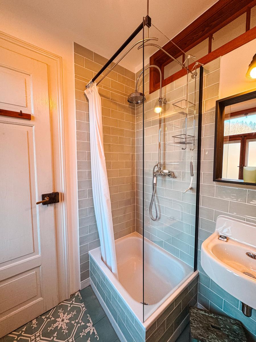 Bad mit Dusche, Waschbecken und Spiegel. Fliesen und Holzrahmen sichtbar.