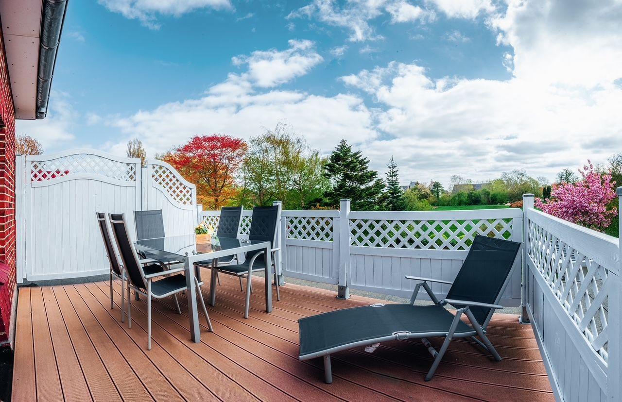 Terrasse mit Tisch, Stühlen und Liegestuhl. Weiße Brüstung, Blick auf Bäume und Himmel.