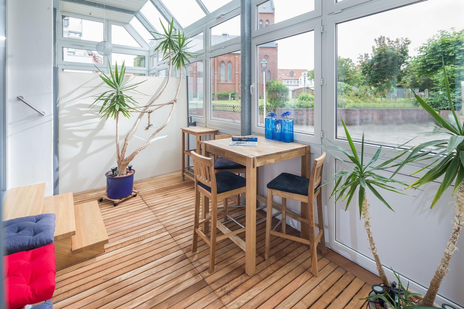 Ein Raum mit Holzboden, Tisch, Stühle und Pflanzen. Große Fenster bieten Blick auf Außenbereich.